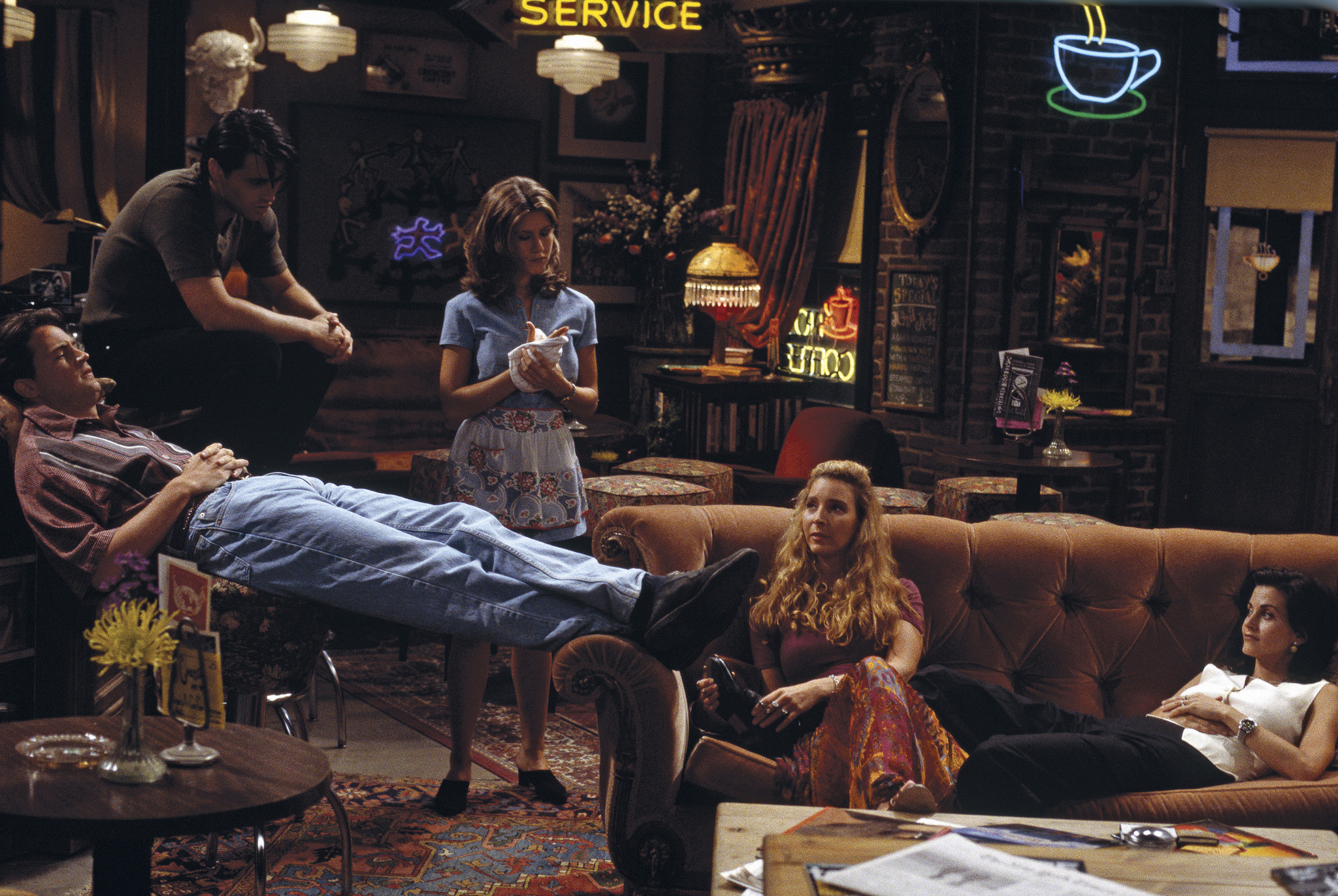 Chandler sits across a chair and couch while Phoebe sits on the couch next to monica lounging across the couch and joey sitting on the bar and Rachel standing in the background